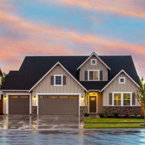 Two-story suburban home with dark grey asphalt shingle roof. Vertical siding with white trim and brown shutters. Rainy evening